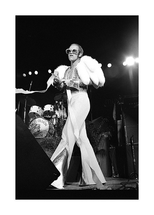  – Mustavalkoinen valokuva laulaja Elton Johnista lavalla yllään valkoinen haalari ja aurinkolasit