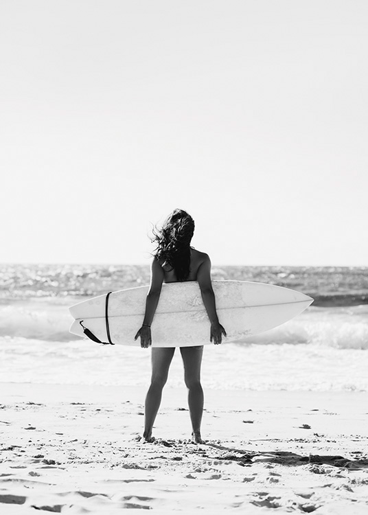  – Mustavalkoinen valokuva tytöstä pitelemässä surffilautaa selkänsä takana valtameri taustanaan