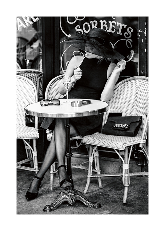  – Mustavalkoinen valokuva naisesta, joka istuu ulkoilmakahvilassa hattu päässään ja sytyttää savuketta